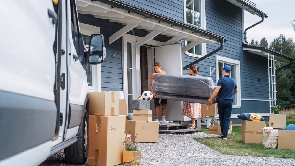 Young Man Helping A Delivery Service Worker To Bring A Modern Couch Into Their New House